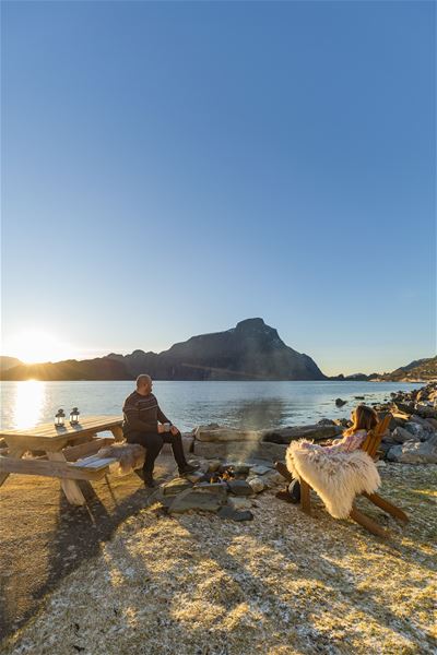 Two people relax by a small campfire near a calm lake, with a picnic table, rocks, and distant mountains at sunset at the Seahouse at Hornelen Panorama in Bremanger..