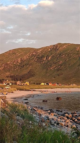 Kystlandskap med sandstrand, steinete fjøre og stille sjø, med hus og grøne fjell under skyer i kveldssol i Hoddevik..