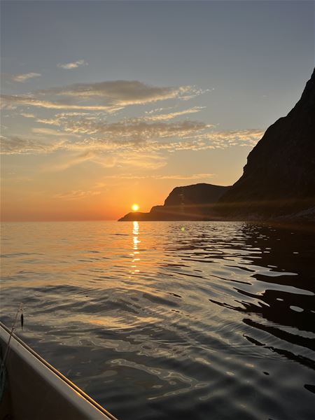 Solnedgang over roleg sjø med gyllent spegelbilete, skyer i himmelen og mørke klipper langs høgre side i Hoddevik..