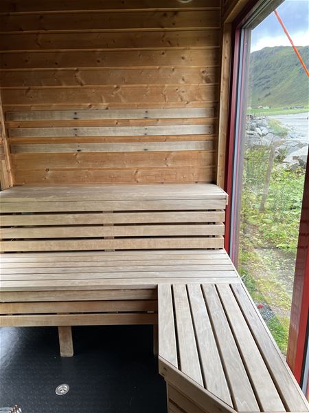 Innvendig rom med trebastu-benk og panelvegg, stort vindauge med utsyn mot steinstrand og fjell i dagslys i Hoddevik..