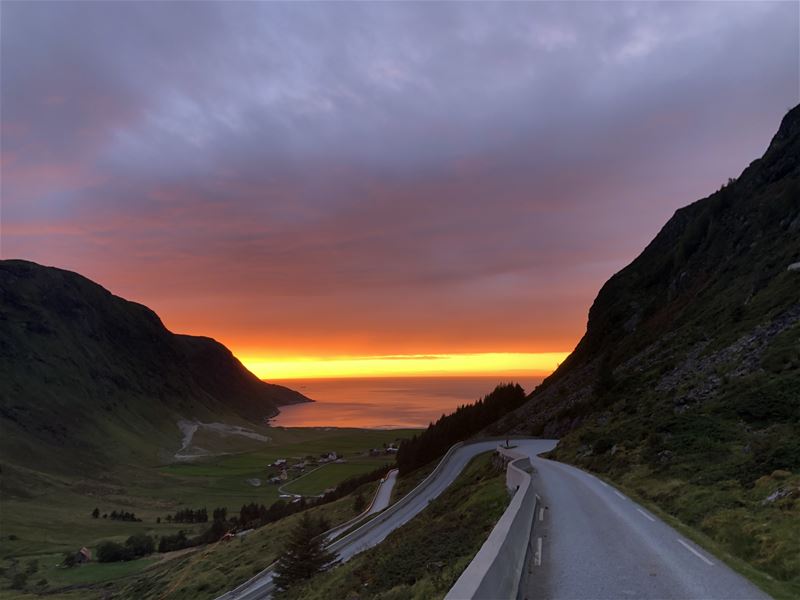 Svingete fjellveg i Hoddevik langs bratt skrent med utsikt til grøne jorder og havet under ein glødande solnedgang..