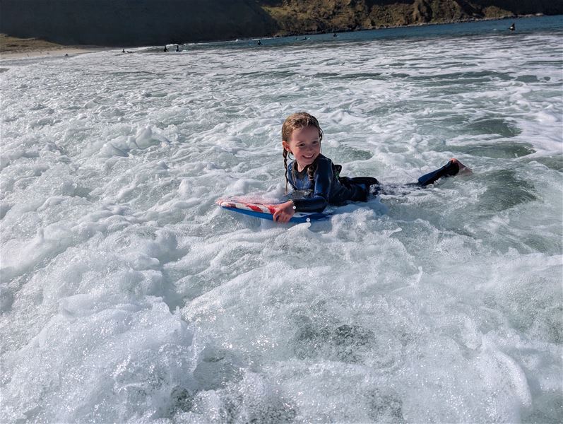 Eit barn i våtdrakt ligg på eit bodyboard i skummande bølgjer nær stranda, med klippekyst i bakgrunnen..