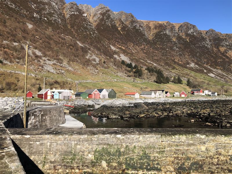 Små naust og hus ved ei steinete fjordstrand, med bratte fjell og blå himmel i bakgrunnen..
