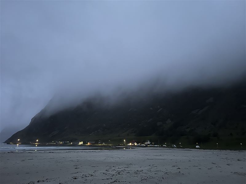 Tåkete kveldslandskap med sandstrand i framgrunnen og ei kystbygd med lys ved foten av mørke fjell i Hoddevik..