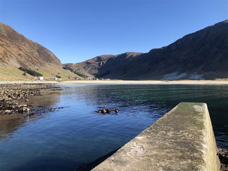 Utsikt over roleg fjord med grunne steinar, strand og bratte fjell på begge sider under klår blå himmel i Nordfjord..