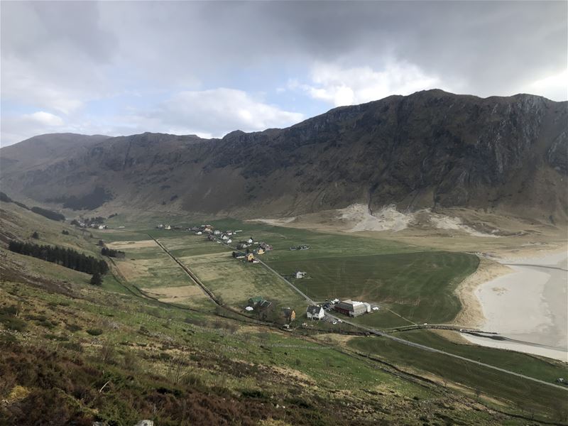 Panoramautsikt over grøne jorder og spreidde gardshus i ei dalbotn, med bratte fjell og skya himmel i Nordfjord..
