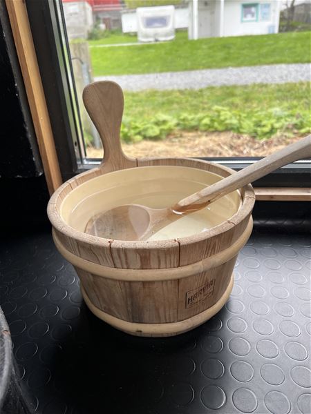 Ei trebøtte fylt med vatn med ei treøse inni står på eit svart golv ved eit vindauge med gras ute..