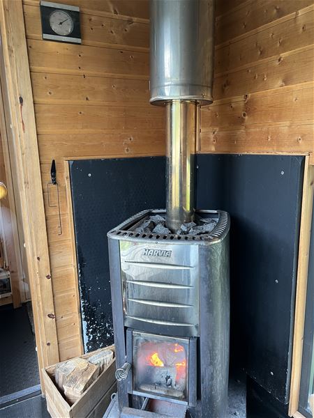 Vedfyrt badstuomn i metall med steinar og synleg flamme, plassert mot trepanel og svart veggvern i Hoddevik i Nordfjord..