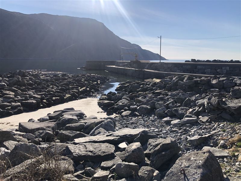 Steinete strand og grunne vassrenner mot ei molo ved stille sjø, med fjell i bakgrunnen og solstråler..