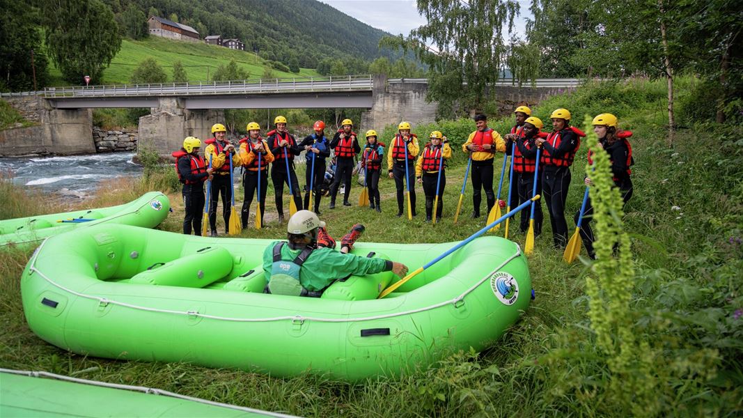 Gruppe med raftingutstyr samlet rundt en grønn flåte ved elvebredden, med bro og dal i bakgrunnen.