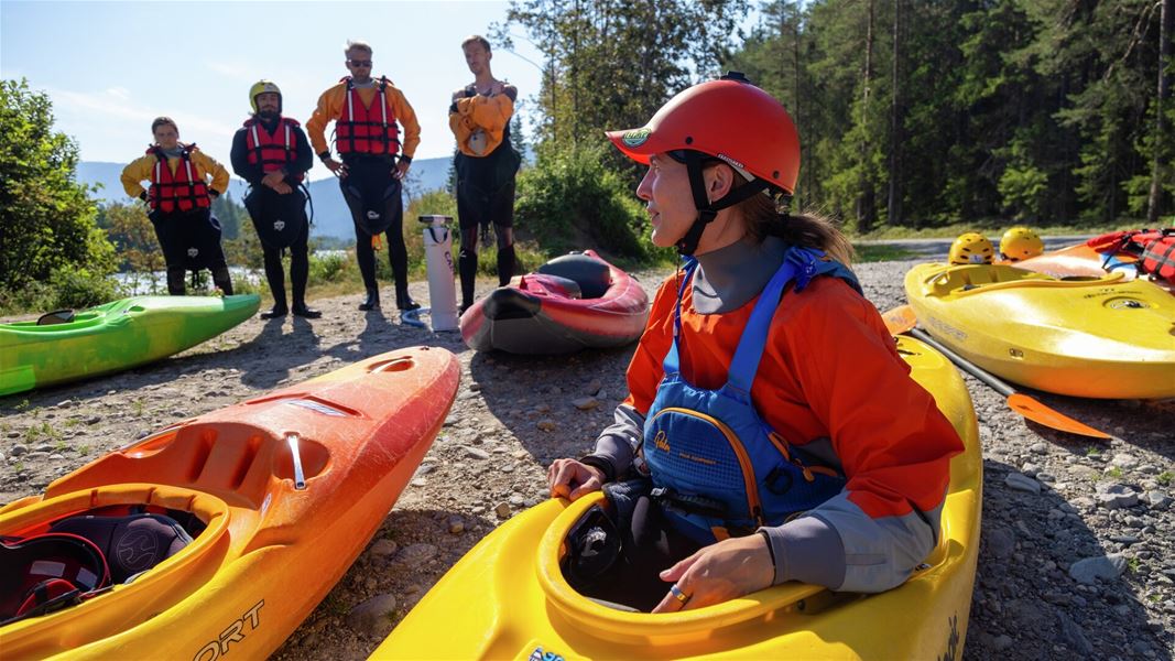 Gruppe med kajakker samlet på elvebredden, med deltakere iført hjelm, redningsvest og padleutstyr.