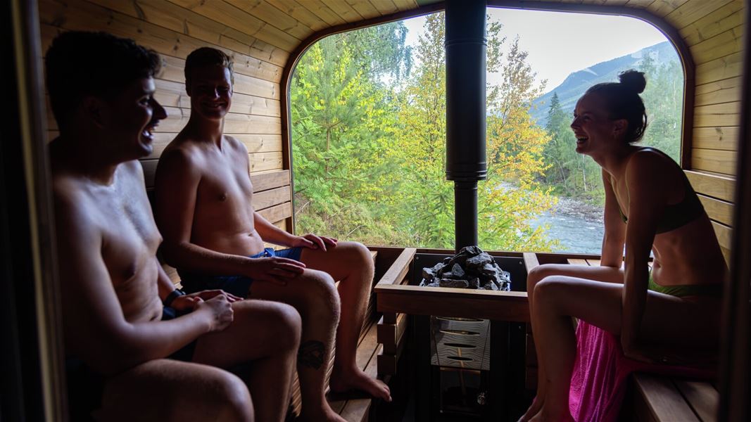Three people sitting inside a sauna with a wood-burning stove and a large window facing forest and river.