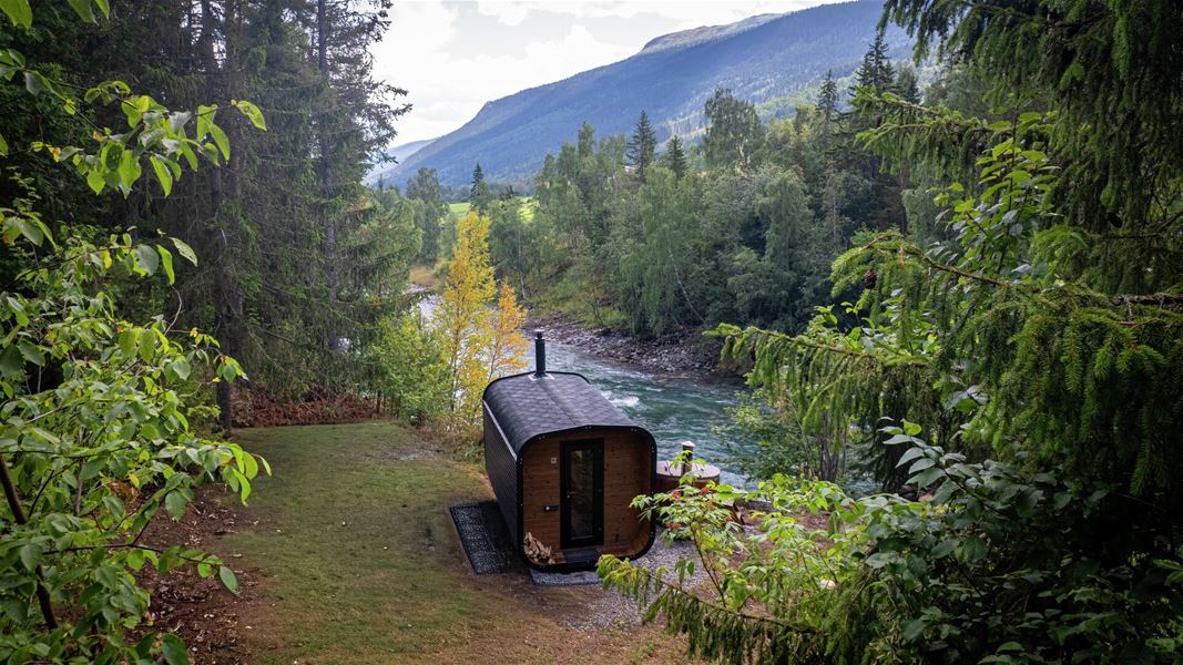 Wood-fired sauna located at the edge of a forest beside a river, with valley and mountains in the background.
