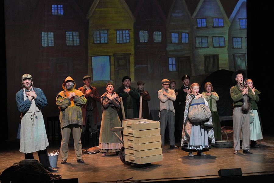Group of actors in period costumes stand on a theater stage before a painted town backdrop, with stacked crates and a cart.