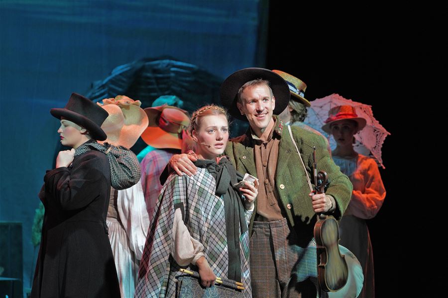 Group of performers in period costumes and hats stand on a lit stage, with one person holding a violin.
