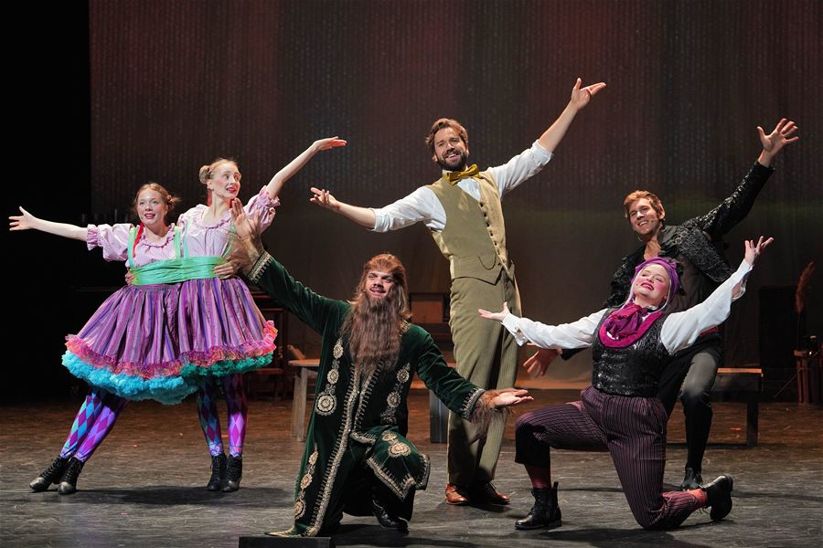 Six performers on a stage hold outstretched arms in a dramatic pose, wearing colorful costumes under warm lighting.