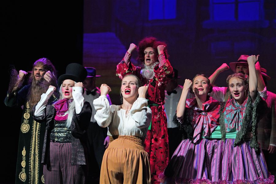 Group of performers in colorful period costumes on a lit stage, raising arms in a synchronized pose.