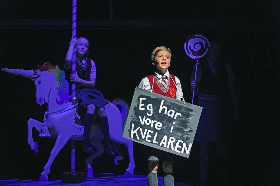 A child performer in a school uniform holds a painted sign on a dark stage with purple lighting and a carousel horse.