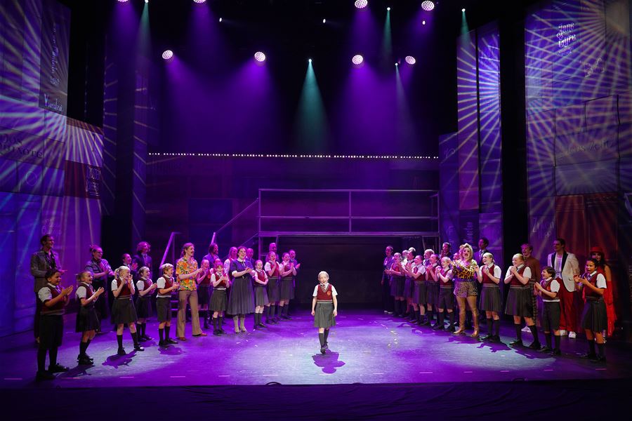 Large cast stands in two lines applauding as a child in a school uniform stands center stage under purple spotlights.