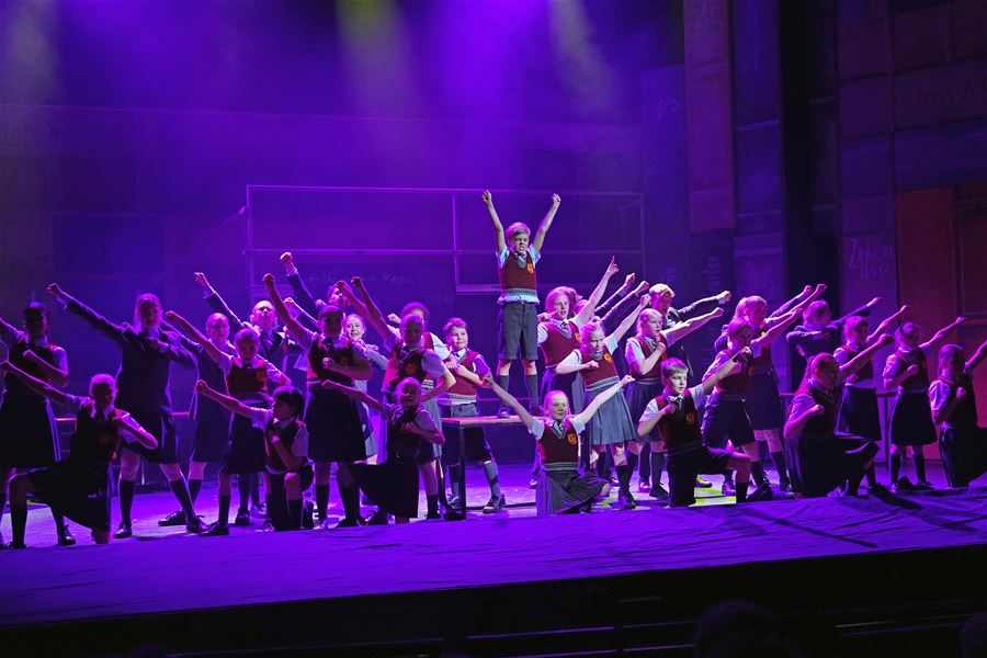 Large group of performers in school-style outfits pose on a stage with raised arms under purple lighting and haze.