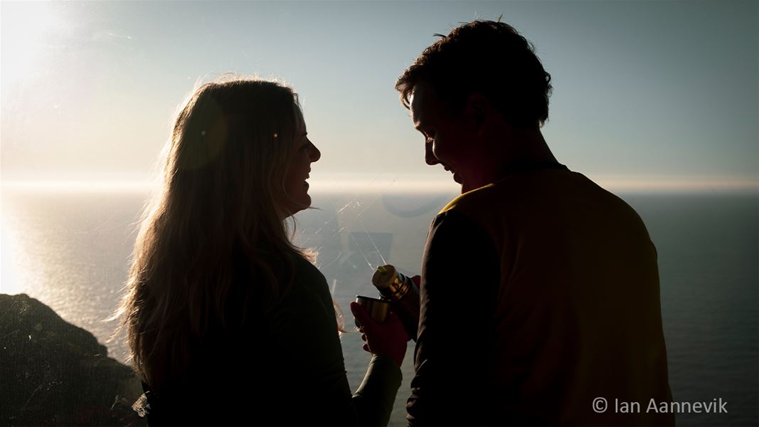 Silhouetted couple facing each other by the sea at sunset, holding a thermos and cup near a coastal overlook..