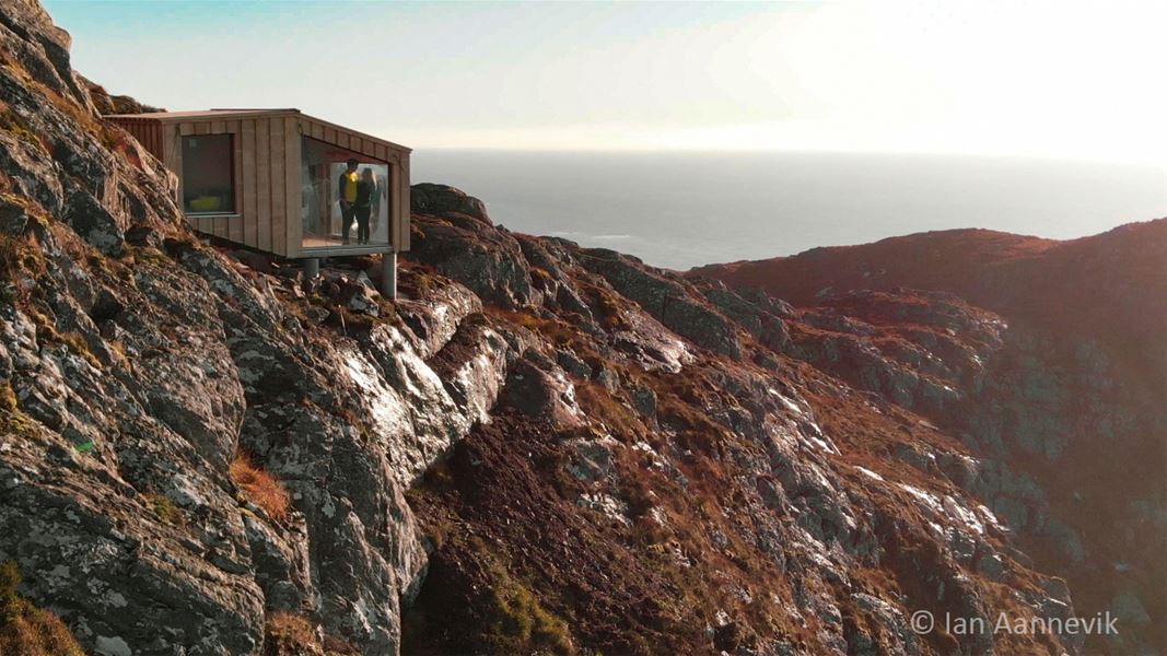 Small modern cabin perched on rugged rocks above the sea, with two people standing by a large window in warm light..
