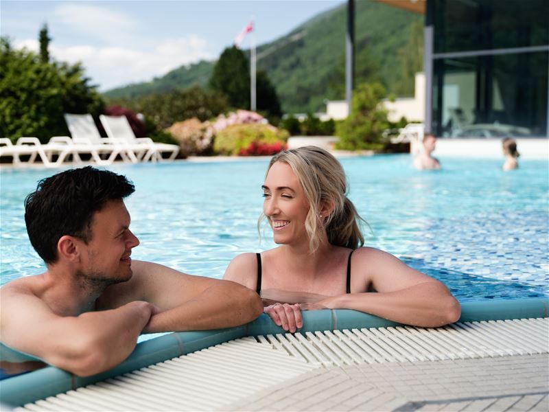 Two people lean on the edge of a blue outdoor pool, with lounge chairs, greenery, and mountains behind..