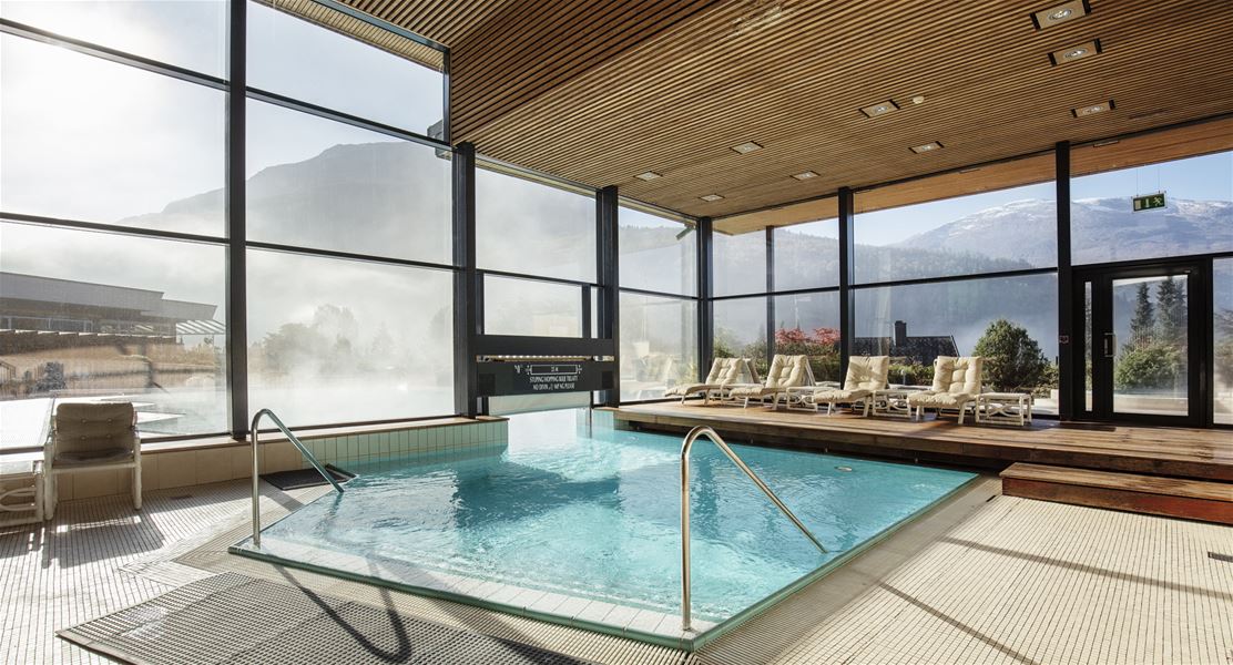Sunlit indoor pool with metal handrails, lounge chairs on a wooden deck, and large windows facing misty mountains at Hotel Alexandra..
