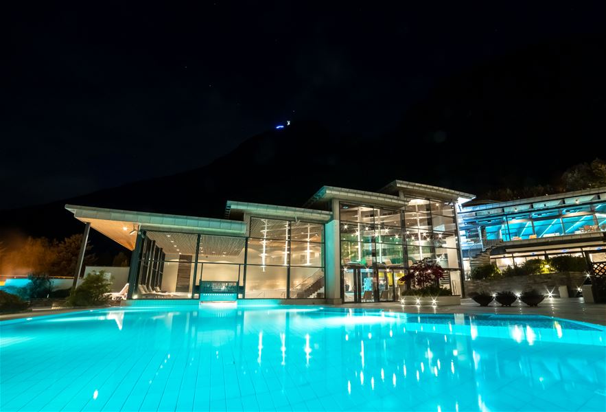 Illuminated outdoor swimming pool in front of a modern glass building at night, with hills silhouetted in the background..