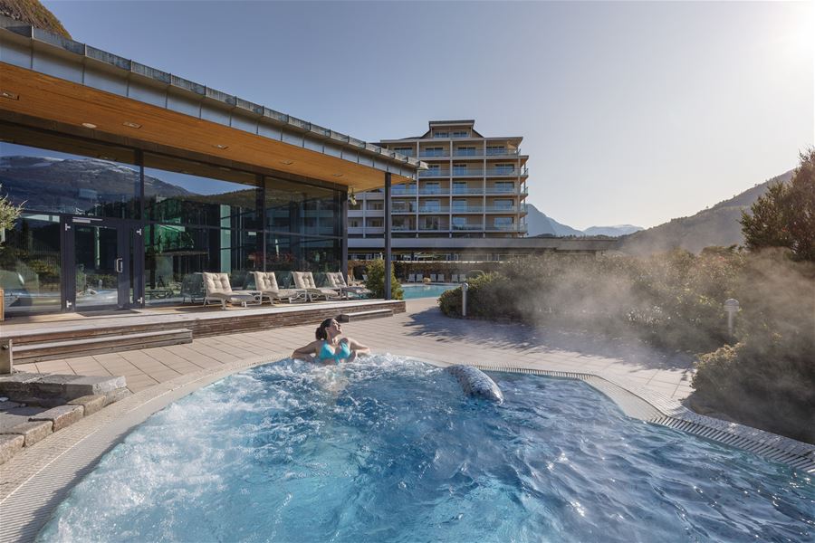 Person relaxing in a bubbling outdoor hot tub beside a modern glass building, with mountains and steam in sunlight..