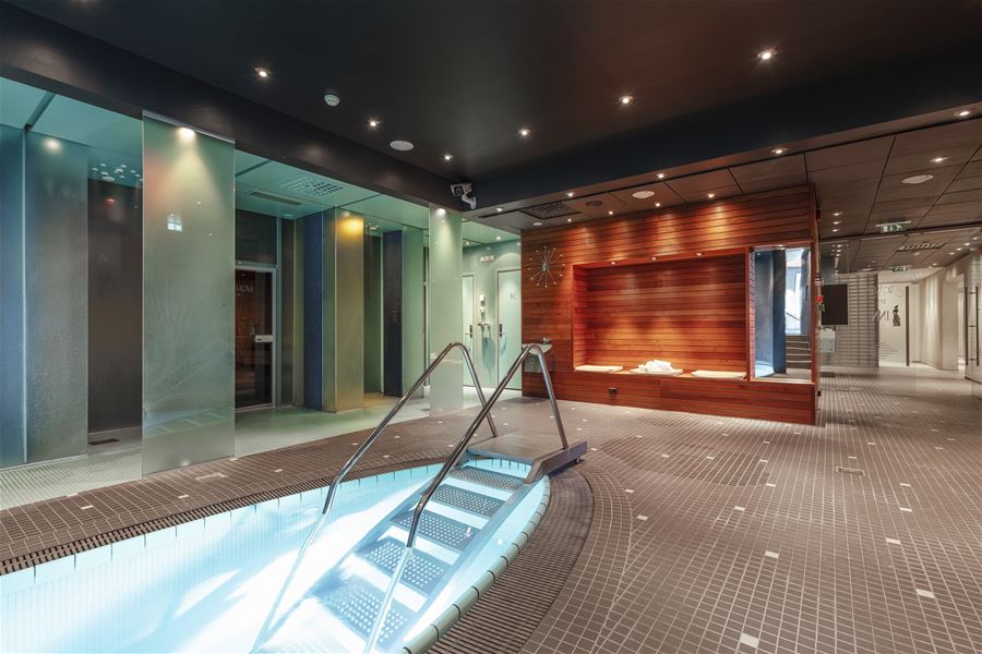 Indoor spa pool edge with metal handrails and lit steps, beside frosted glass walls and a wood bench alcove at Alexandra Spa..