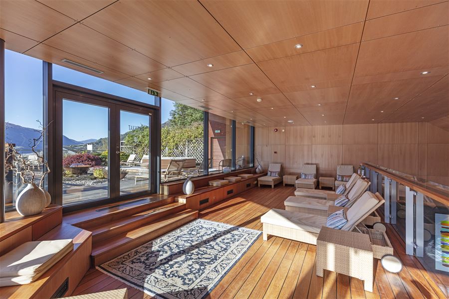 Bright wooden lounge with reclining chairs, rug, and large glass doors opening to a terrace and distant mountains at Alexandra Spa..