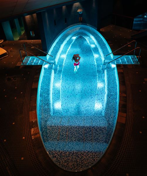 Overhead view of an oval indoor pool with blue lighting, mosaic tiles, side steps, and a lone swimmer at Alexandra Spa.