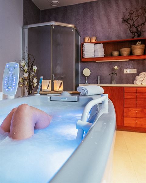Person soaking in a bubbling jacuzzi tub in a spa-like bathroom with towels, candles, shelves, and a shower stall at Hotel Alexandra..