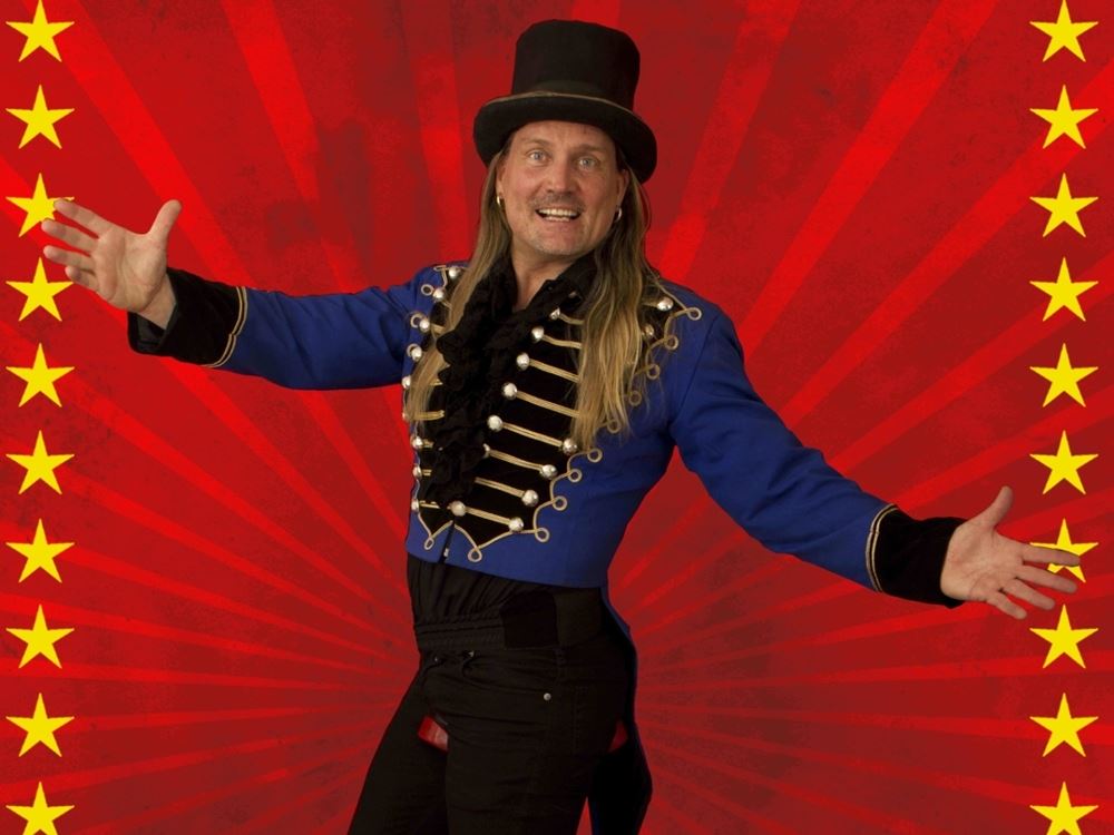 En person i blå cirkusjacka och hög hatt står med utsträckta armar mot en röd bakgrund med stjärnor..