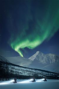 Three snowmobiles drive while the Aurora forks a path in the sky during the night