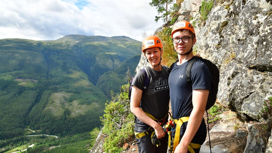 Two people in climbing gear standing by a cliff with mountain views.