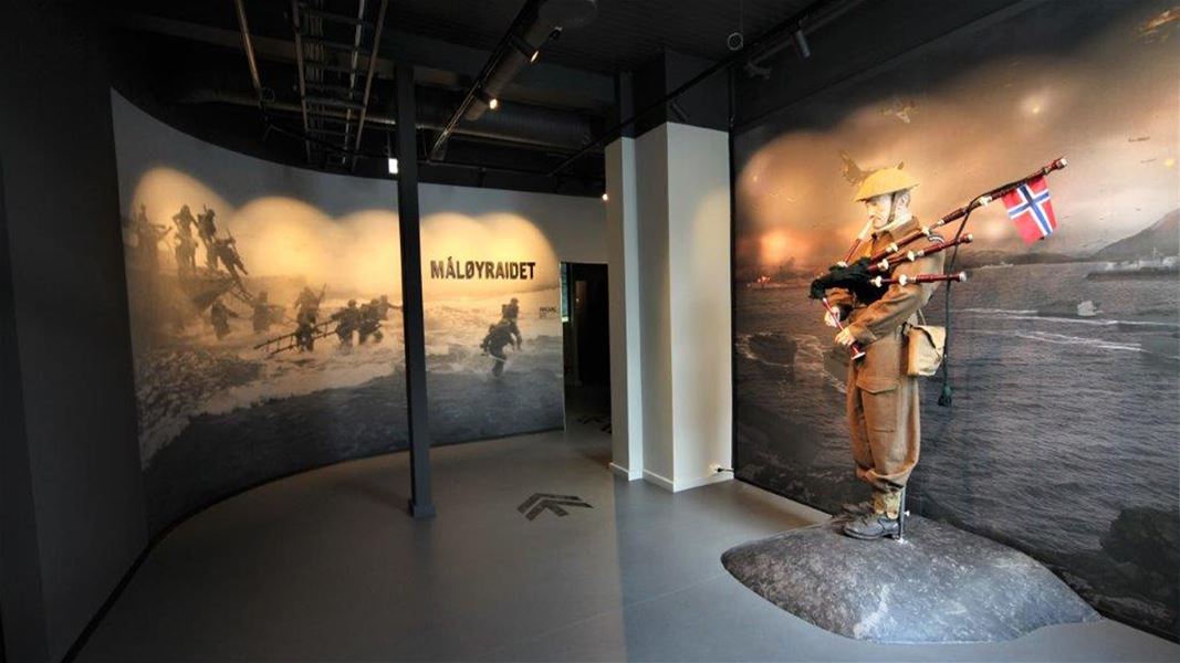 Museumsrom med store veggbilete av krigsscener og ein uniformert soldatfigur med sekkepipe og norsk flagg..