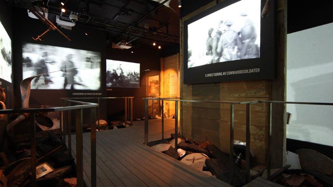 Mørkt museumsrom med gangbru og fleire skjermar som viser svart-kvitt krigsfilm og dokumentasjon på veggane..