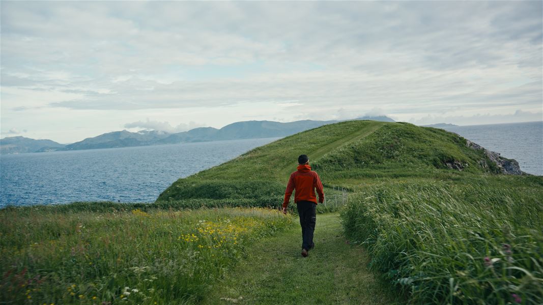 A person in a red jacket walks along a grassy trail on a coastal hill, with ocean and distant mountains under clouds..