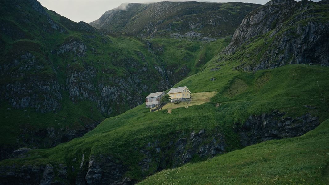 A small yellow house and outbuilding sit on a grassy hillside, surrounded by steep rocky green mountains under clouds..