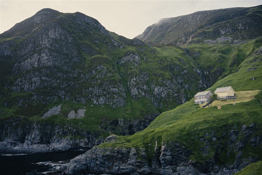 A lone yellow house sits on a grassy cliff above dark water, backed by steep rocky mountains and misty sky..