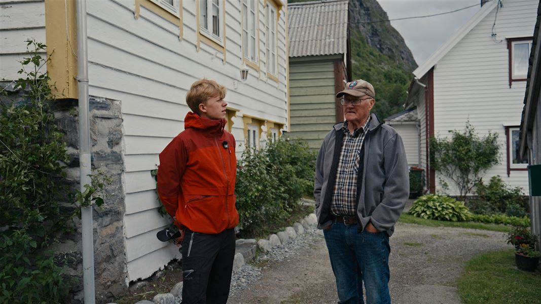 Two men talk in a small village yard beside wooden houses, with shrubs and a rocky hillside in the background..