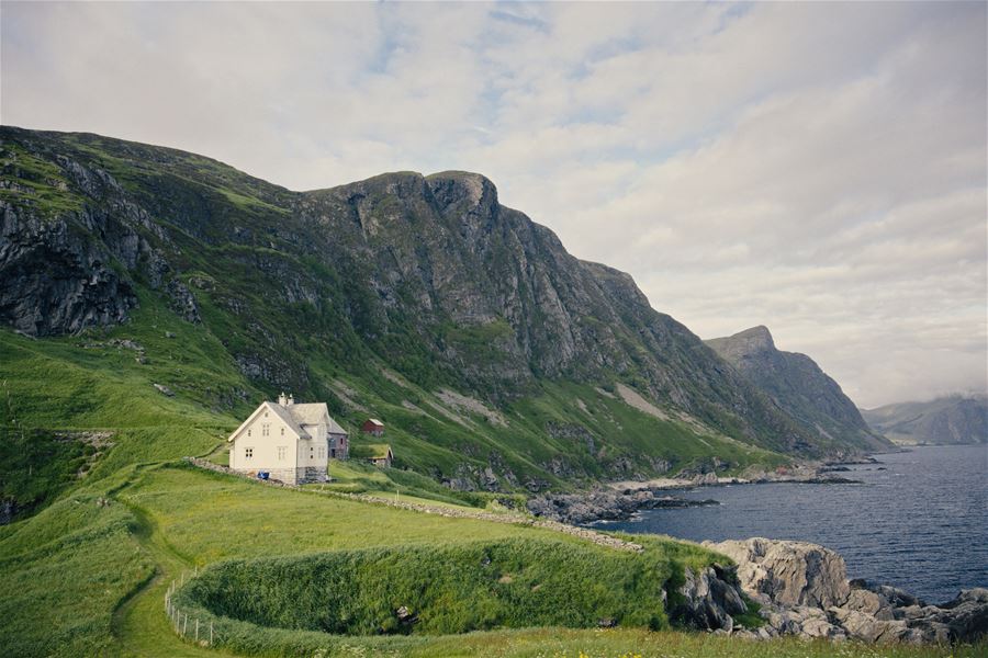 A small white house sits on a grassy hillside beside rocky coastline, with steep green mountains under a cloudy sky..