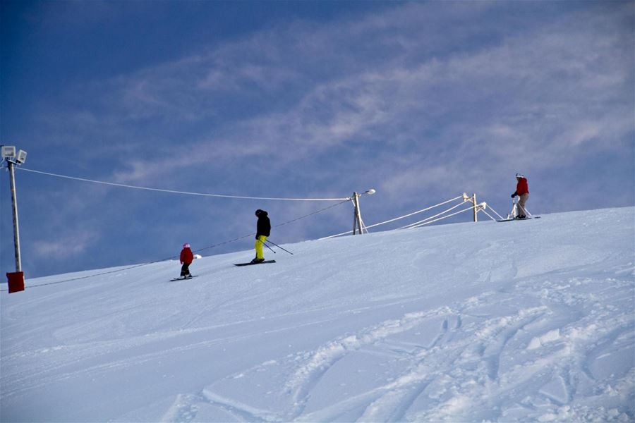 Three skiers on a snowy slope under a cloudy blue sky, with lift cables and poles along the ridgeline..