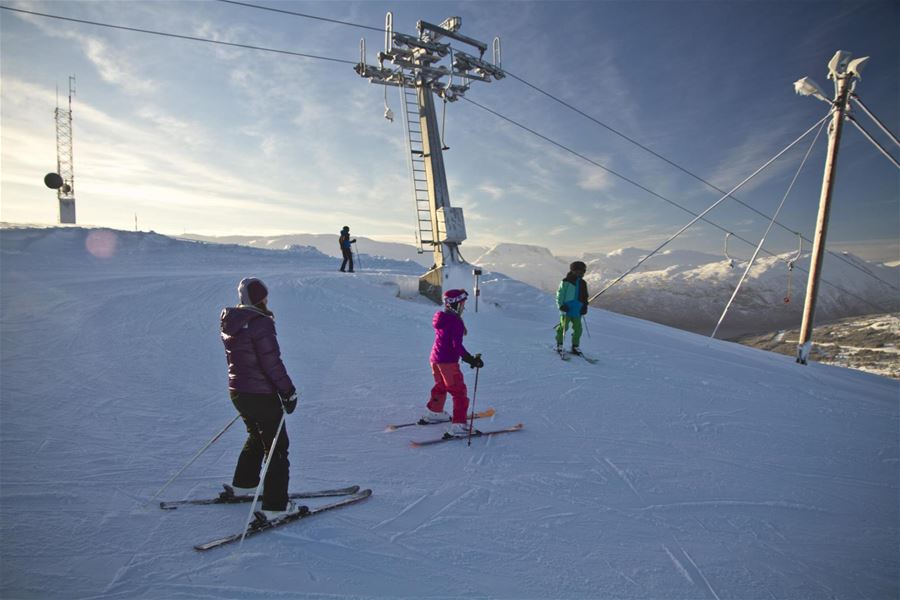 Three skiers stand on a snowy slope near a lift tower, with distant mountains under a clear sky..