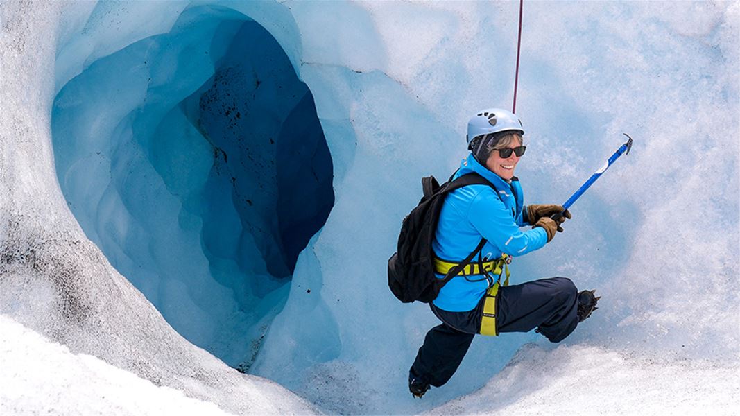 A climber wearing helmet and backpack uses an ice axe on a snowy glacier beside a deep blue crevasse opening..