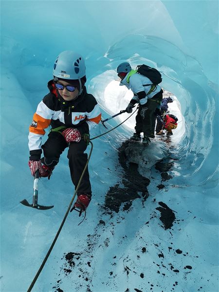 Roped ice climbers wearing helmets use ice axes and crampons while moving through a blue glacier tunnel..