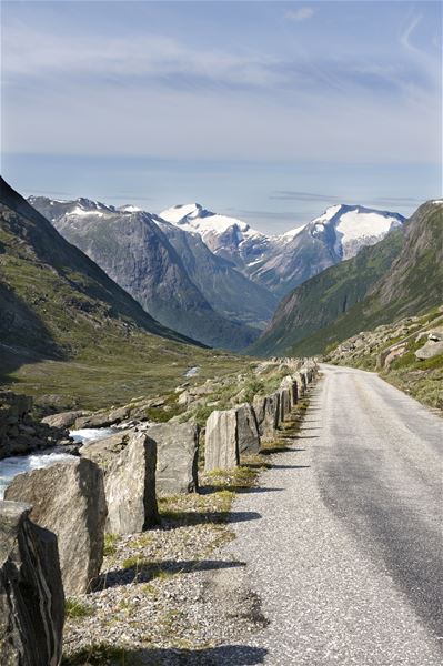 Narrow paved road with stone barriers winds through a green mountain valley toward snow-capped peaks under blue sky..