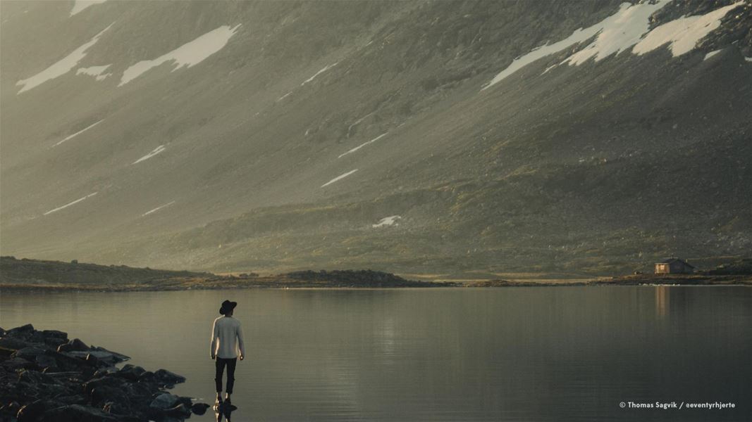 A person stands on rocks at a calm lake, facing a large mountain slope with patches of snow and soft light..